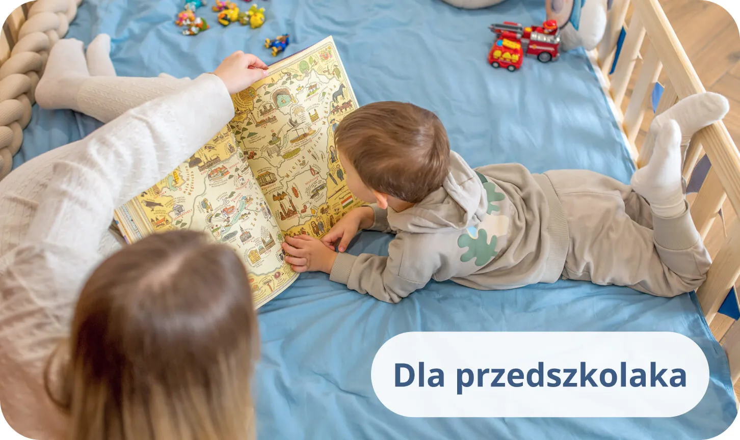 Mały chłopiec z mamą czytający kolorową książkę na niebieskiej pościeli w sosnowym łóżku.